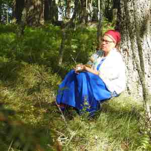 Itkuvirsien itkijä Emilia Kallonen istuu puun juurella karjalaisnaisen asussa 