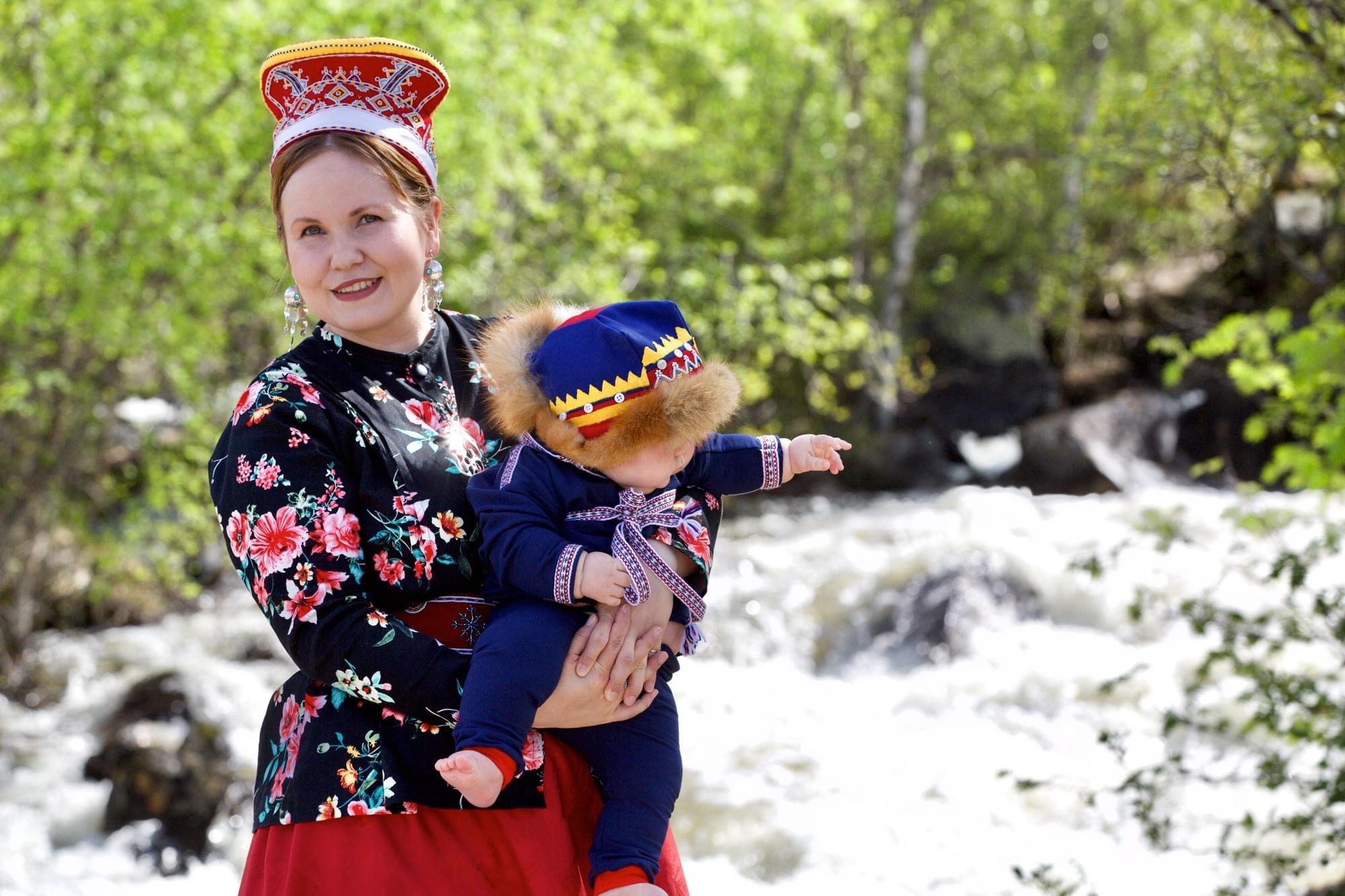 Anna Lumikivi kolttasaamelaisen asussa lapsi sylissään joen rannalla