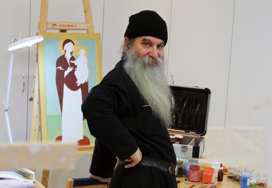 Pitkäpartainen ja mustaan viittaan pukeutunut ikonimaalari isä Luukas katsoo kameraan puolitekoinen ikoni taustallaan Valamon luostarin ikonimaalausluokassa