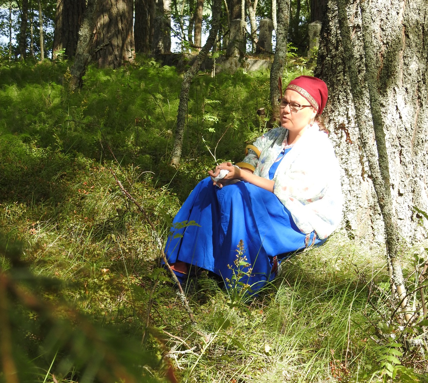 Itkuvirsien itkijä Emilia Kallonen istuu puun juurella karjalaisnaisen asussa